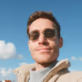 Smiling man wearing sunglasses and a beige jacket against a clear blue sky.